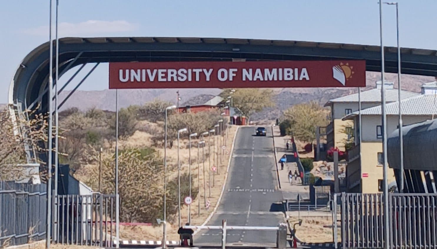 Eine asphaltierte Straße, die unter einem gewölbten Schild mit der Aufschrift University of Namibia hindurchführt.