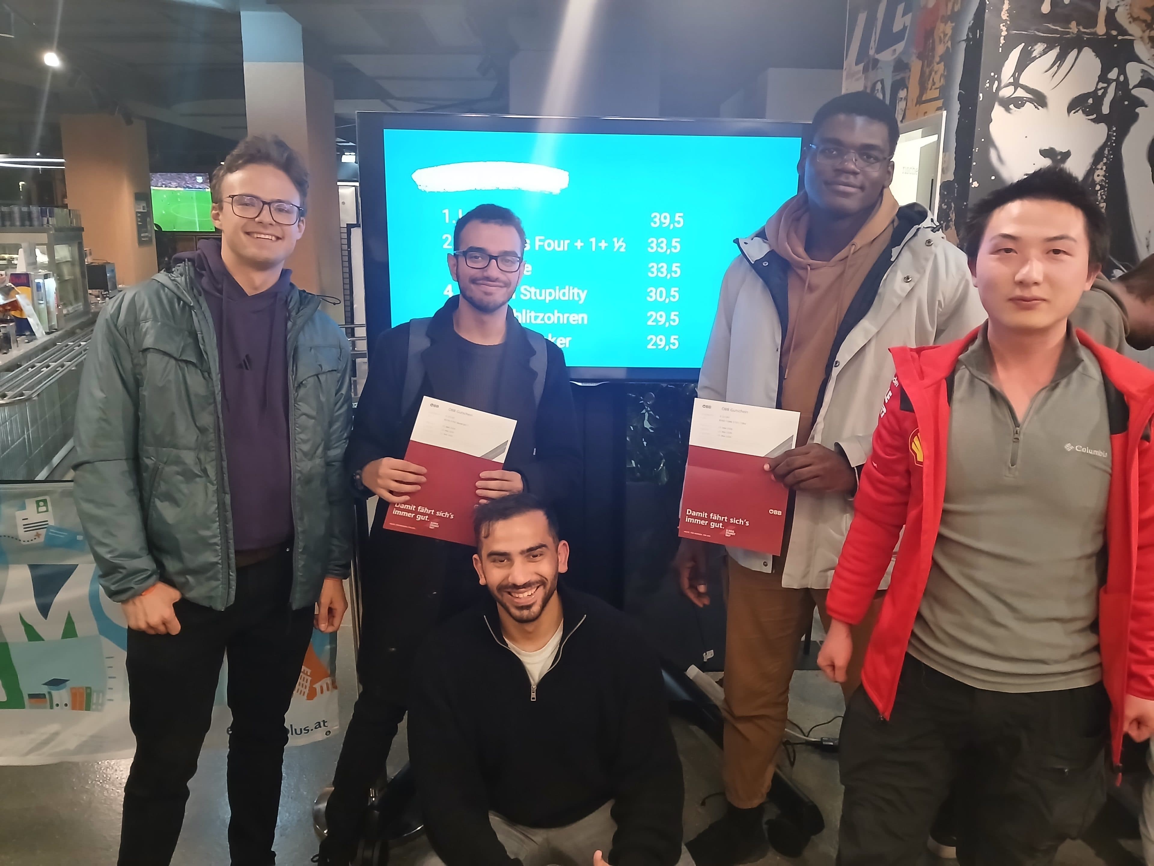 Fünf junge Männer posieren für ein Foto vor einem blauen Hintergrund, auf dem eine Anzeigetafel zu sehen ist; zwei der Männer halten rote ÖBB-Unternehmensmappen in der Hand.