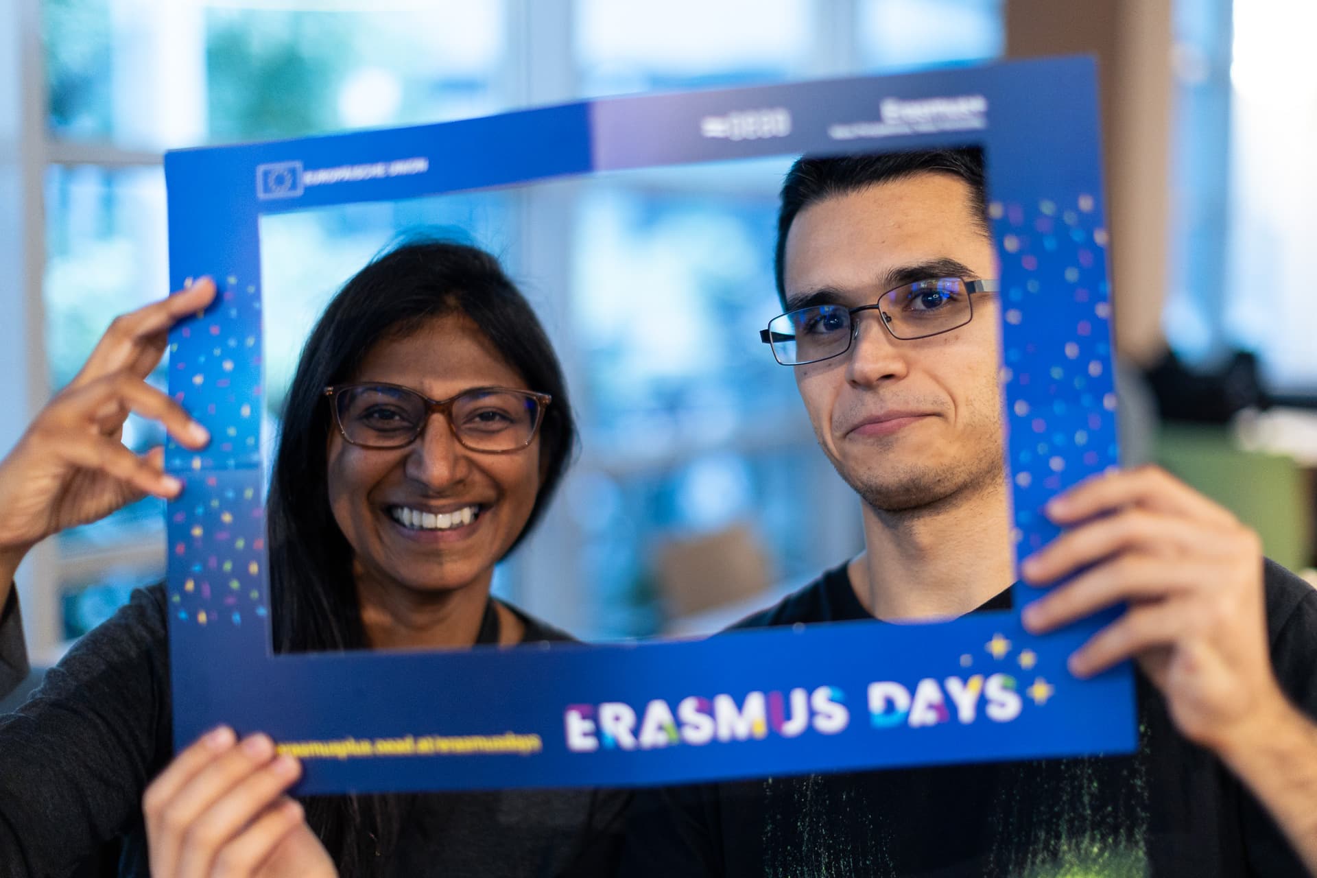 Ein Mann und eine Frau, die lächeln, während sie einen blauen Bilderrahmen mit dem Erasmus Days-Logo vor ihr Gesicht halten.