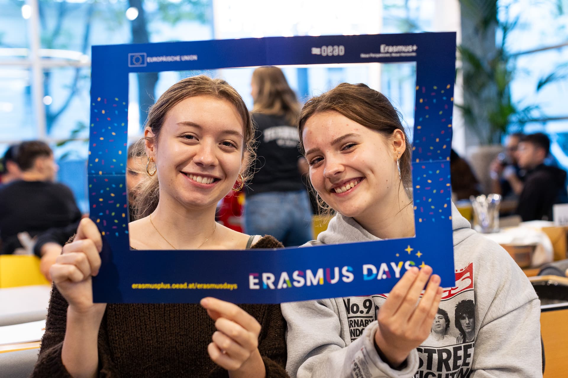 Zwei junge Frauen lächeln, während sie einen blauen Erasmus Days-Fotorahmen vor ihr Gesicht halten.