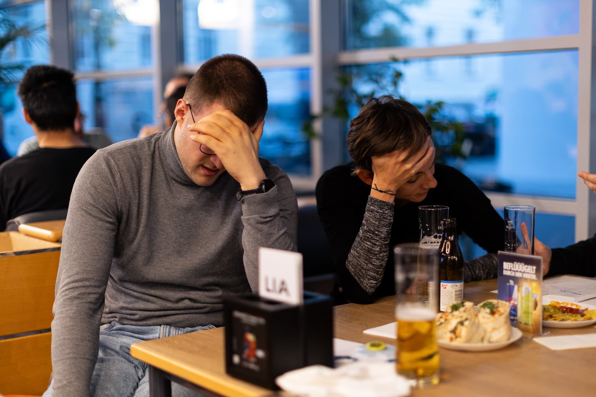 Zwei Menschen sitzen an einem Tisch, haben den Kopf in die Hände gestützt und sehen frustriert aus.
