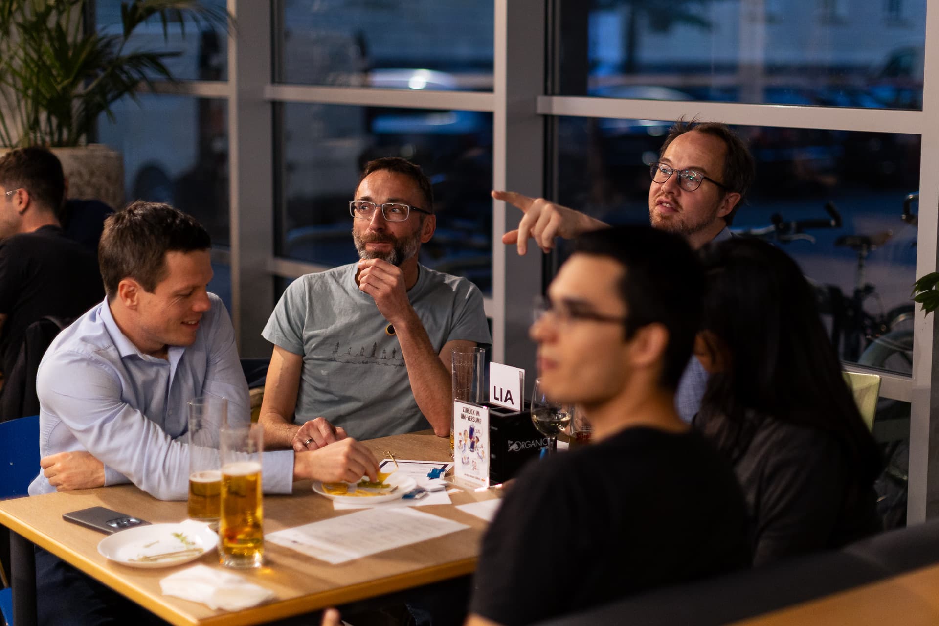 Eine Gruppe von Menschen sitzt an einem Tisch in einem Restaurant; ein Mann zeigt auf ein Fenster, während die anderen zuschauen.