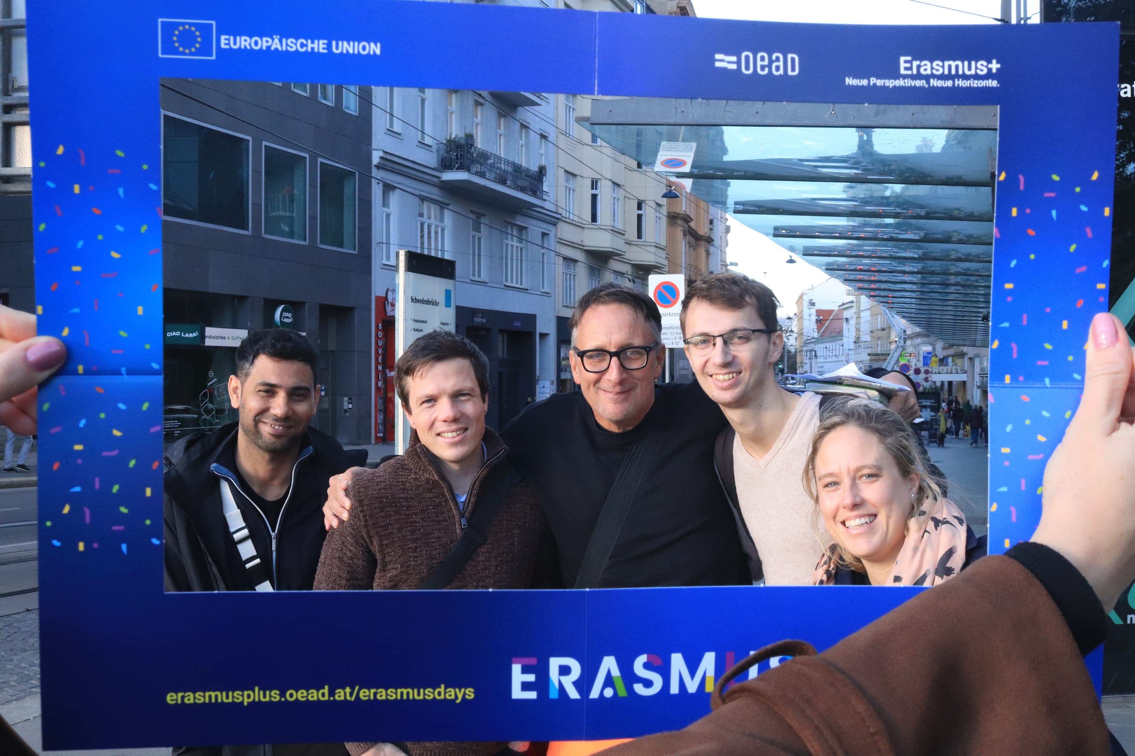 Fünf Personen posieren gemeinsam hinter einem Fotorahmen mit dem Erasmus+-Logo auf einer Straße in der Stadt.