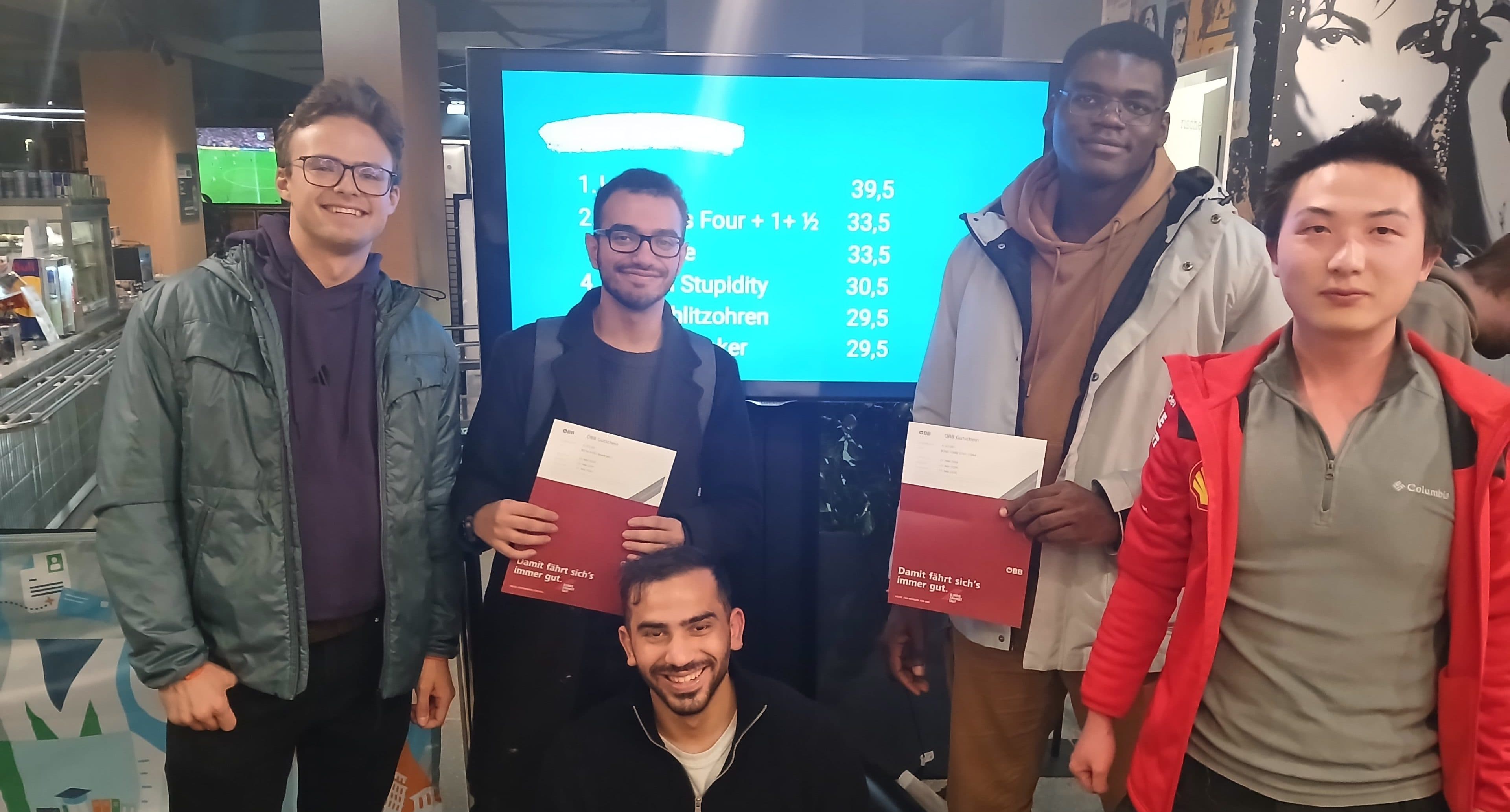 Vier junge Männer posieren für ein Foto vor einem Bildschirm, auf dem eine Liste mit Namen und Punkteständen zu sehen ist; zwei von ihnen halten ÖBB-Broschüren in der Hand.